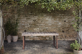 The nkuku Oso Wooden Dining Table stands on a dark stone patio by an aged brick wall, with green vines overhead and potted plants beside it, creating a serene and natural outdoor setting.