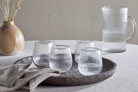 A textured tablecloth holds a round tray with nkuku’s Otuba Tumbler - Clear (Set of 4) made from recycled ribbed glass, next to a folded beige napkin. In the background sit a matching ribbed glass pitcher and a ceramic vase with dried stems.