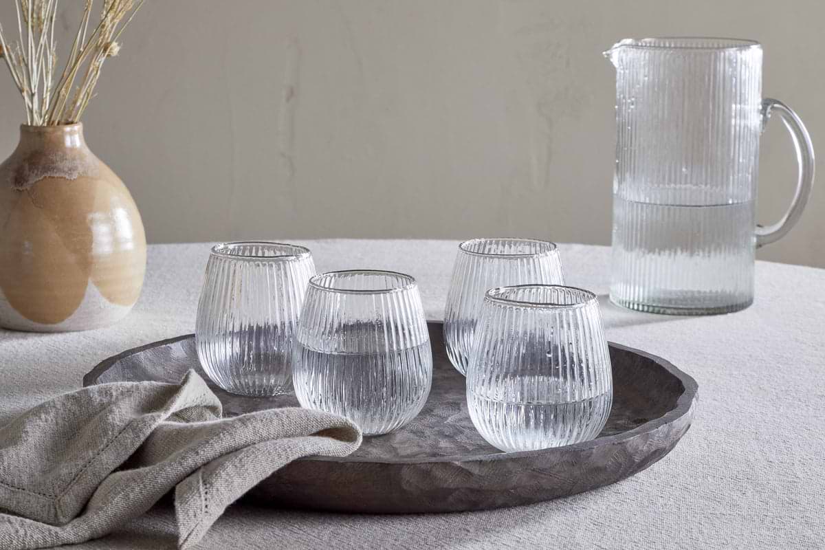 A textured tablecloth holds a round tray with nkuku’s Otuba Tumbler - Clear (Set of 4) made from recycled ribbed glass, next to a folded beige napkin. In the background sit a matching ribbed glass pitcher and a ceramic vase with dried stems.