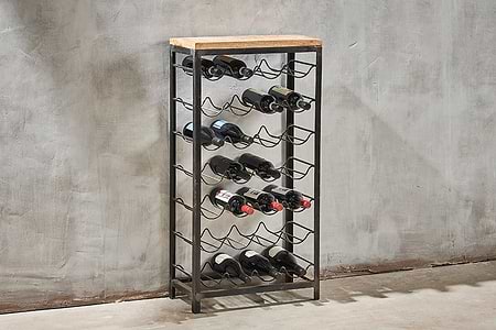 The nkuku Obra Industrial Wine Rack - Mango Wood & Iron - Large features five iron shelves for horizontal bottle storage and a mango wood top, its modern design accentuated by a gray concrete wall and minimalist setting.