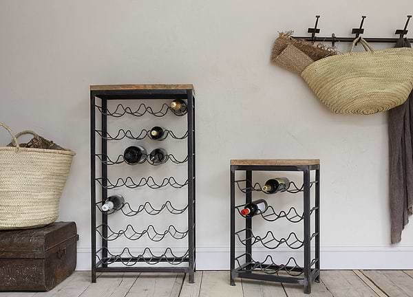 Two nkuku Obra Industrial Wine Racks—crafted from mango wood and iron in a small size—stand on a wooden floor against a light wall, holding several bottles; wicker baskets and a brown blanket hang above vintage chests and extra storage.