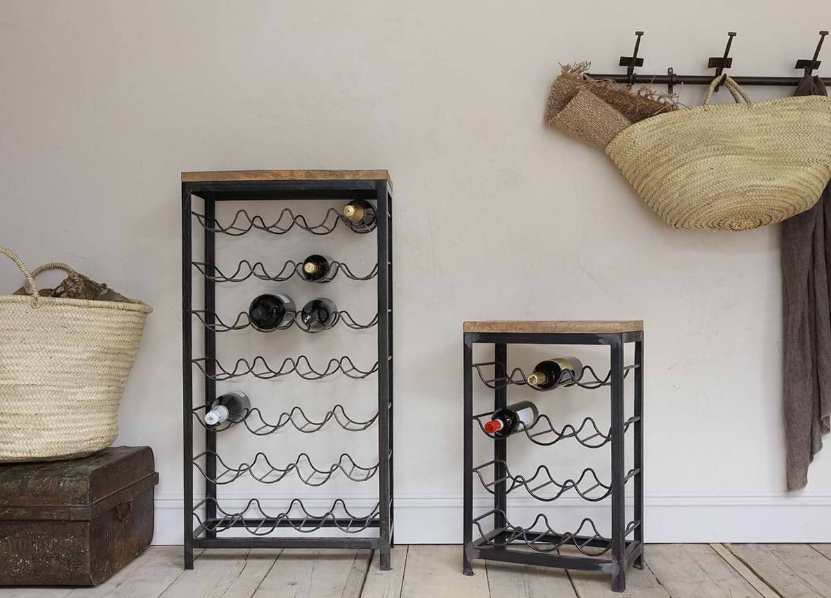 Two nkuku Obra Industrial Wine Racks—crafted from mango wood and iron in a small size—stand on a wooden floor against a light wall, holding several bottles; wicker baskets and a brown blanket hang above vintage chests and extra storage.