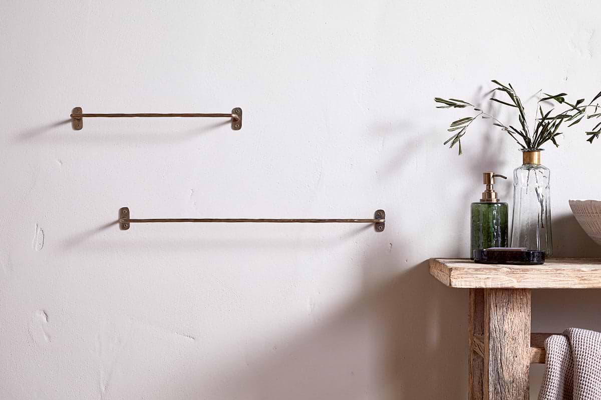 A minimalist bathroom features two nkuku Olani Bathroom Hanging Rails in antique brass on a white wall. To the right, a rustic wood shelf holds a glass bottle with olive branches, a green soap dispenser, and neatly folded towels.