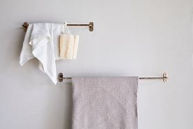 Two nkuku Olani Bathroom Hanging Rail racks in antique brass are mounted on a light bathroom wall. The top rack holds a white towel, washcloth, and beige bath sponge; the lower rack displays a textured beige towel. The wall is unadorned.