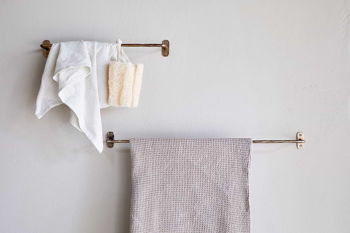 Two nkuku Olani Bathroom Hanging Rail racks in antique brass are mounted on a light bathroom wall. The top rack holds a white towel, washcloth, and beige bath sponge; the lower rack displays a textured beige towel. The wall is unadorned.