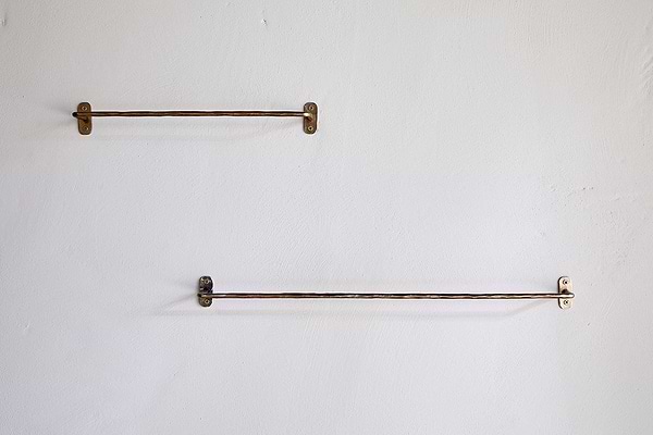Two nkuku Olani Bathroom Hanging Rails in antique brass are mounted on a light bathroom wall; the upper rail is shorter and to the left, while the lower, longer rail is offset to the right, both fixed with visible screws.