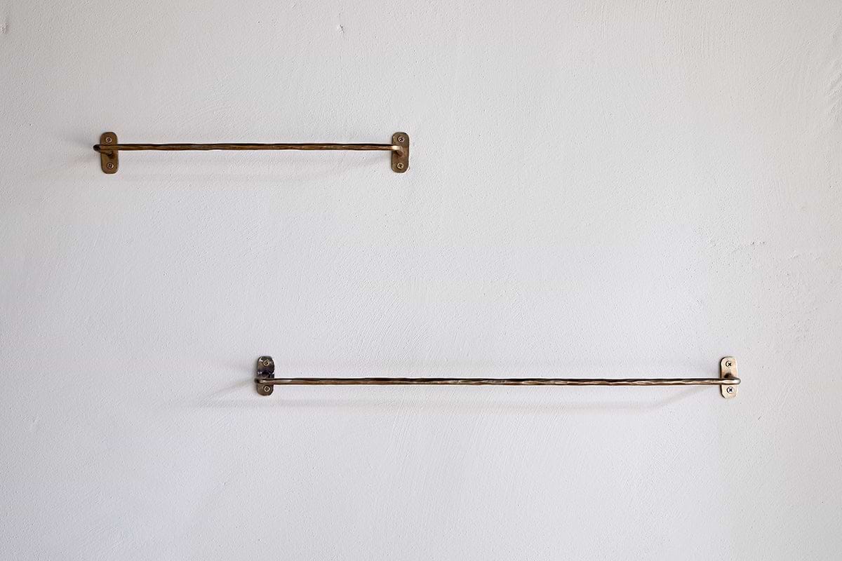 Two nkuku Olani Bathroom Hanging Rails in antique brass are mounted on a light bathroom wall; the upper rail is shorter and to the left, while the lower, longer rail is offset to the right, both fixed with visible screws.