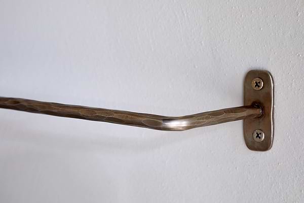 A close-up of an nkuku Olani Bathroom Hanging Rail in antique brass with a hammered texture, mounted on a white wall with a rectangular bracket and two visible screws; the bar is slightly bent, and the wall surface is smooth.