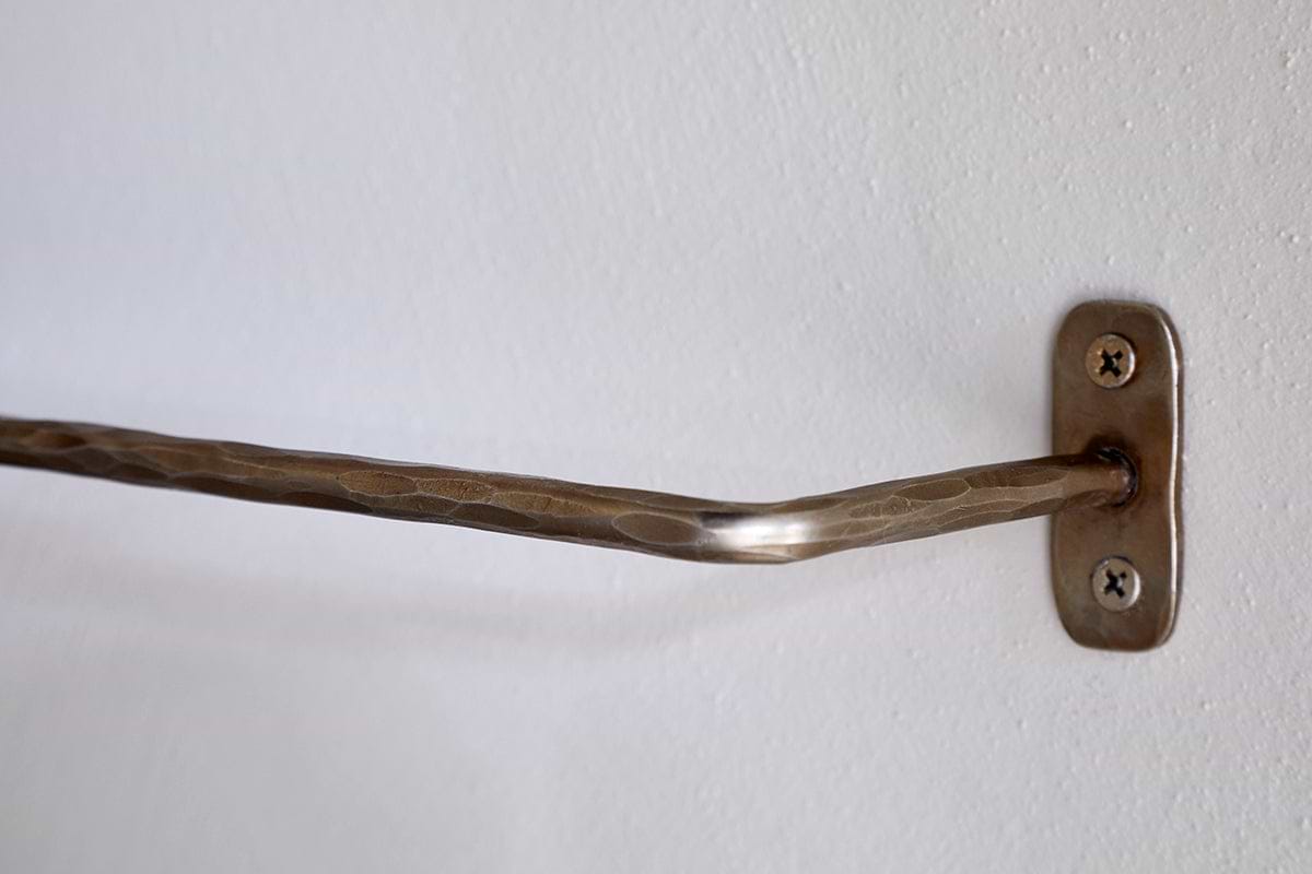 A close-up of an nkuku Olani Bathroom Hanging Rail in antique brass with a hammered texture, mounted on a white wall with a rectangular bracket and two visible screws; the bar is slightly bent, and the wall surface is smooth.
