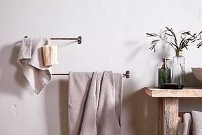 A minimal bathroom scene featuring beige towels on the nkuku Olani Bathroom Hanging Rail in Antique Brass, a loofah, and a rustic wooden shelf with a green glass soap dispenser, olive branches in a vase, and a bowl against a light wall.