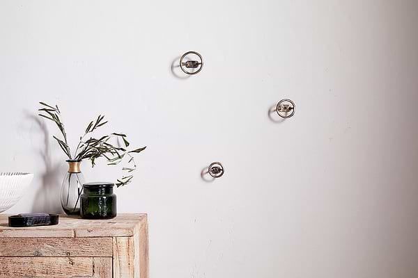 A minimalist scene features a light wooden table with a green glass jar, white bowl, black object, and clear vase with olive branches. On the plain white wall above are three Olani Bathroom Hooks in antique brass by nkuku.