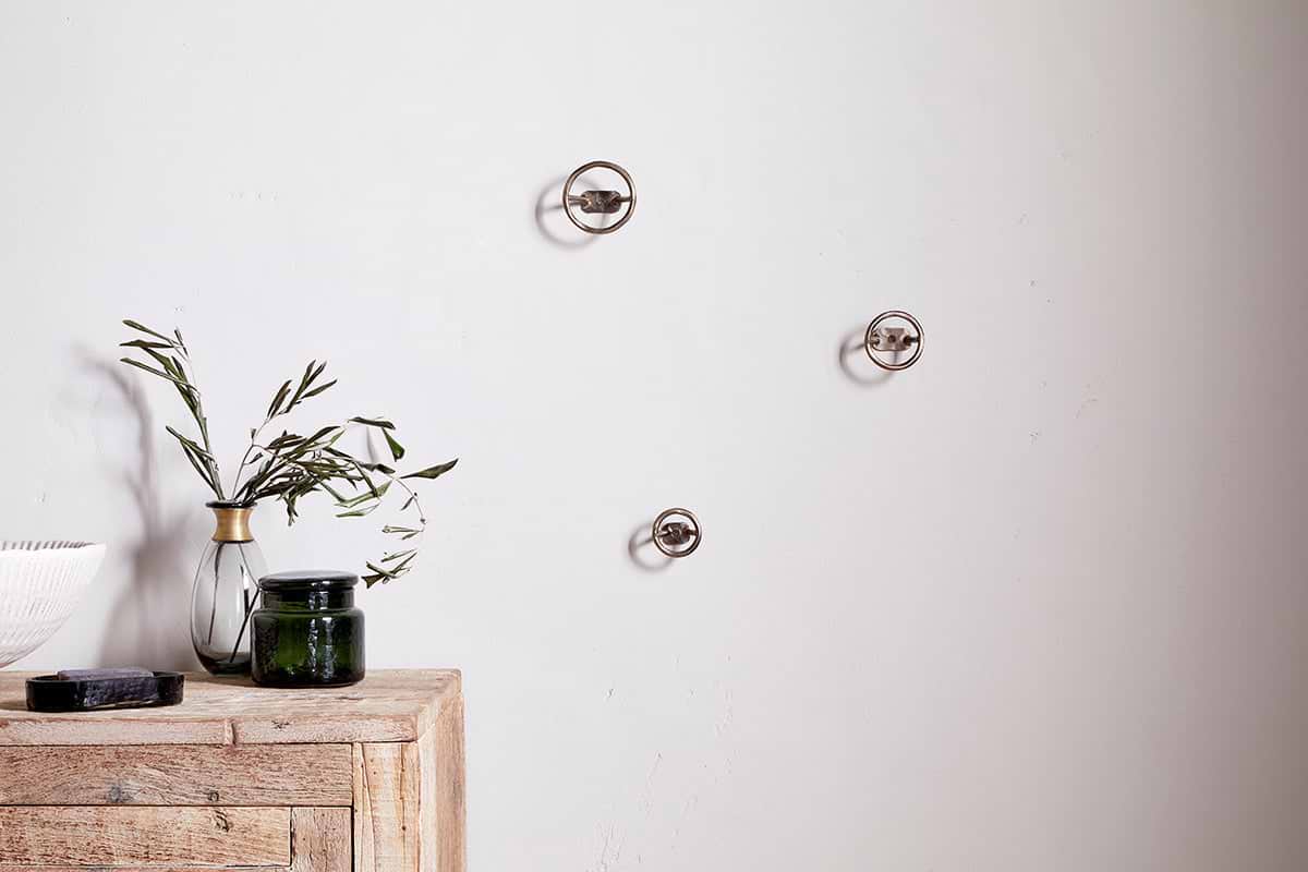 A minimalist scene features a light wooden table with a green glass jar, white bowl, black object, and clear vase with olive branches. On the plain white wall above are three Olani Bathroom Hooks in antique brass by nkuku.