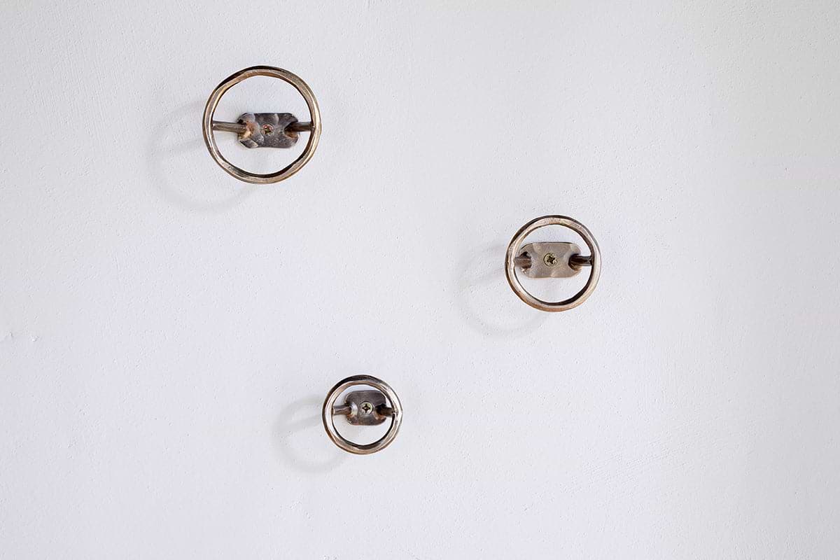 Three Olani Bathroom Hooks by nkuku in antique brass are mounted on a plain white wall in a loose triangular formation, casting soft shadows and creating a subtle, spa-like ambiance.
