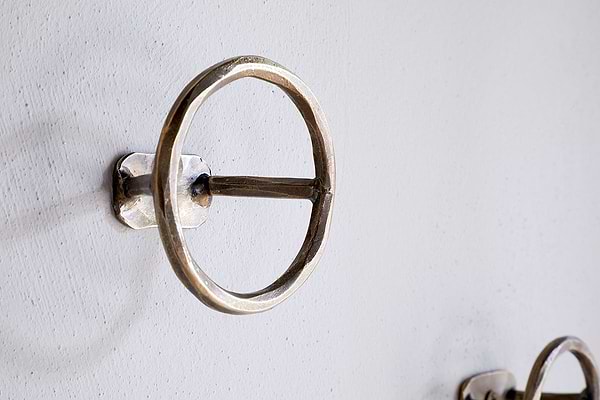 A close-up of an nkuku Olani Bathroom Hook in antique brass, featuring a round design with a horizontal bar, mounted on a white textured wall. A second hook from the set of 3 appears in the background for a spa-like feel.