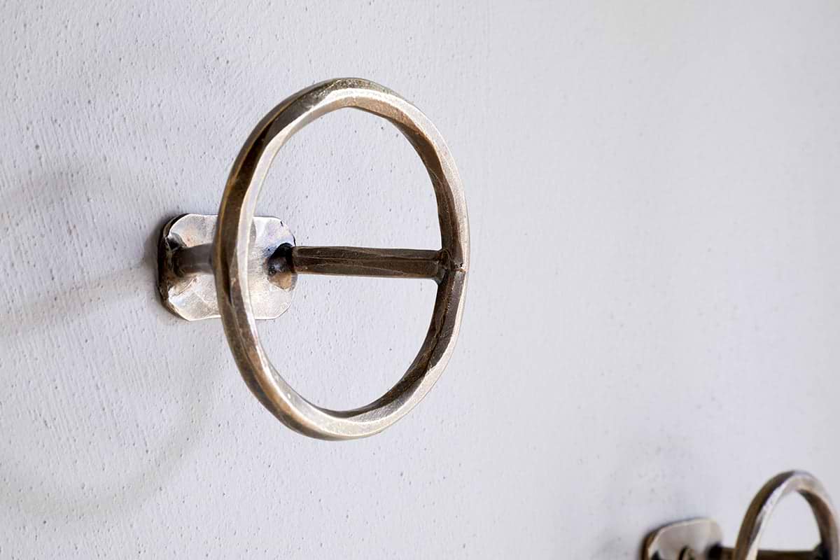A close-up of an nkuku Olani Bathroom Hook in antique brass, featuring a round design with a horizontal bar, mounted on a white textured wall. A second hook from the set of 3 appears in the background for a spa-like feel.