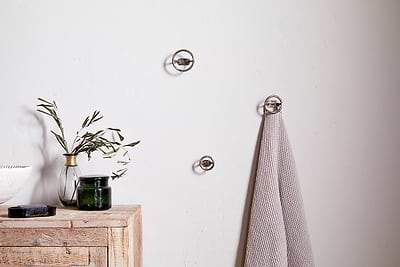 A minimalist spa bathroom features a light wooden cabinet, green branches in a vase, a dark candle, and a gray towel hanging on nkuku's Olani Bathroom Hooks in Antique Brass (Set of 3) against a plain white wall.