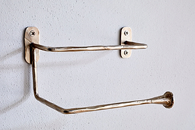 The Olani Toilet Roll Holder by nkuku features a handcrafted, hammered texture in antique brass. Its angular, stainless steel design mounts to a white bathroom wall with two visible screws per plate and stands empty, ready for a new roll.