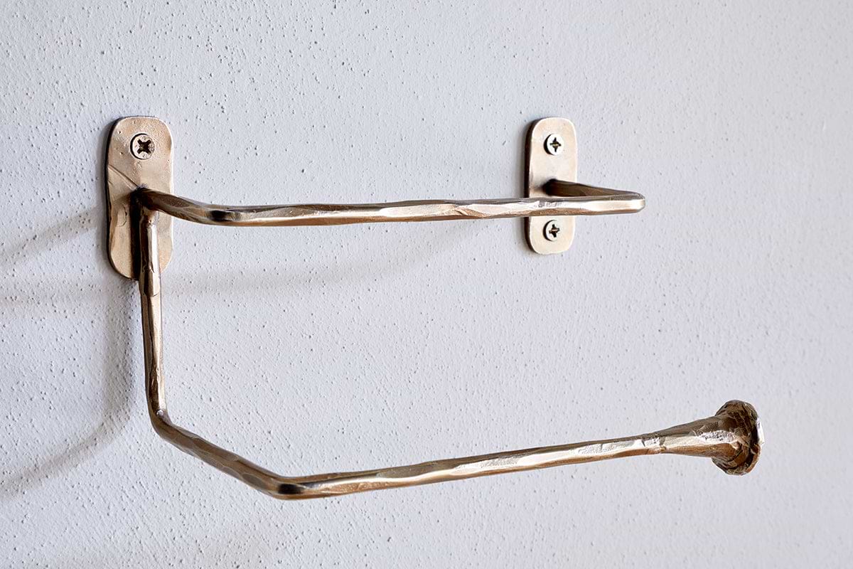 The Olani Toilet Roll Holder by nkuku features a handcrafted, hammered texture in antique brass. Its angular, stainless steel design mounts to a white bathroom wall with two visible screws per plate and stands empty, ready for a new roll.