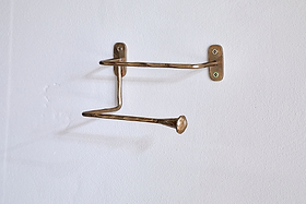 The nkuku Olani Toilet Roll Holder in antique brass features two bent, uneven bars with flared flat ends, hand-forged for a unique look. Wall-mounted with two screws, it casts a subtle shadow against the white bathroom wall.