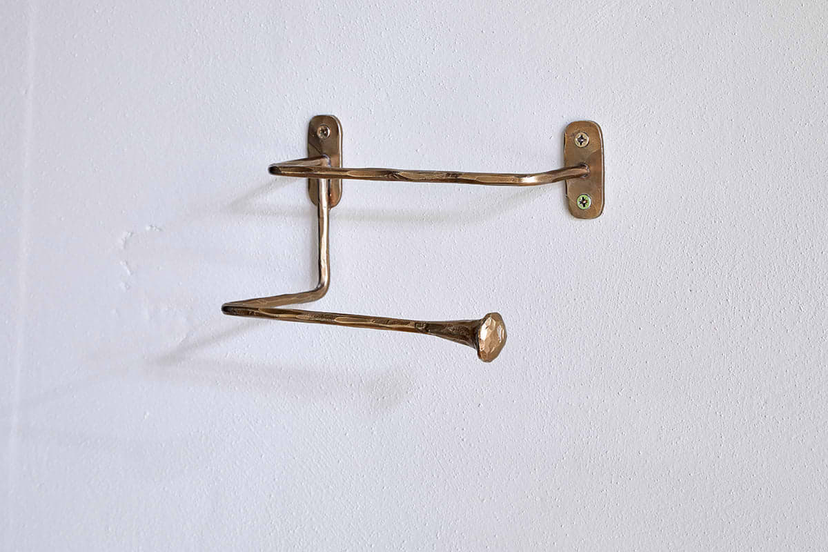 The nkuku Olani Toilet Roll Holder in antique brass features two bent, uneven bars with flared flat ends, hand-forged for a unique look. Wall-mounted with two screws, it casts a subtle shadow against the white bathroom wall.
