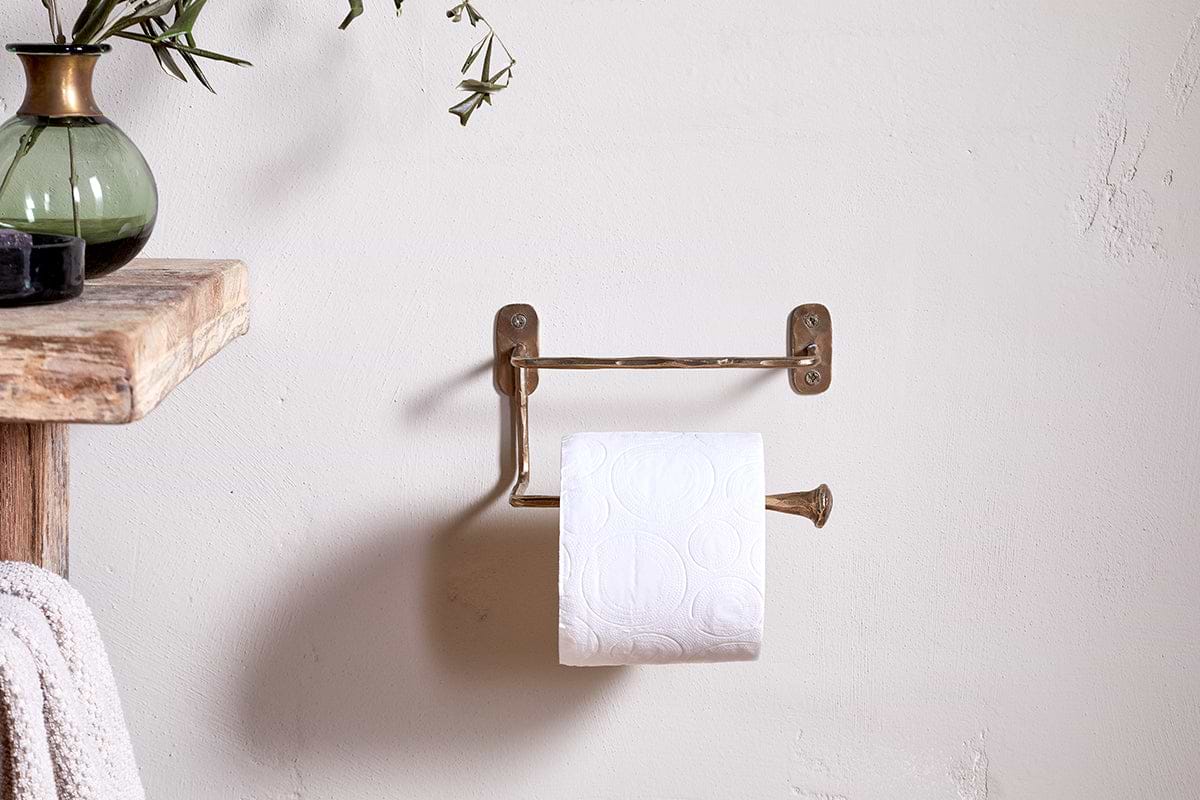 The Olani Toilet Roll Holder in antique brass by nkuku is mounted on a light bathroom wall with a toilet paper roll, beside a wooden shelf displaying a green vase and towel, enhancing the rustic, minimalistic decor.