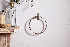 The Olani Towel Ring by nkuku in antique brass is mounted on a white wall. A rustic wooden shelf nearby holds a striped cup with toothbrushes, a small bowl, and a glass bottle with leafy stems for a minimal, natural bathroom look.