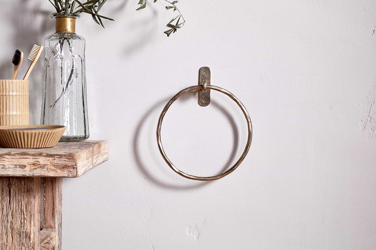 The Olani Towel Ring by nkuku in antique brass is mounted on a white wall. A rustic wooden shelf nearby holds a striped cup with toothbrushes, a small bowl, and a glass bottle with leafy stems for a minimal, natural bathroom look.