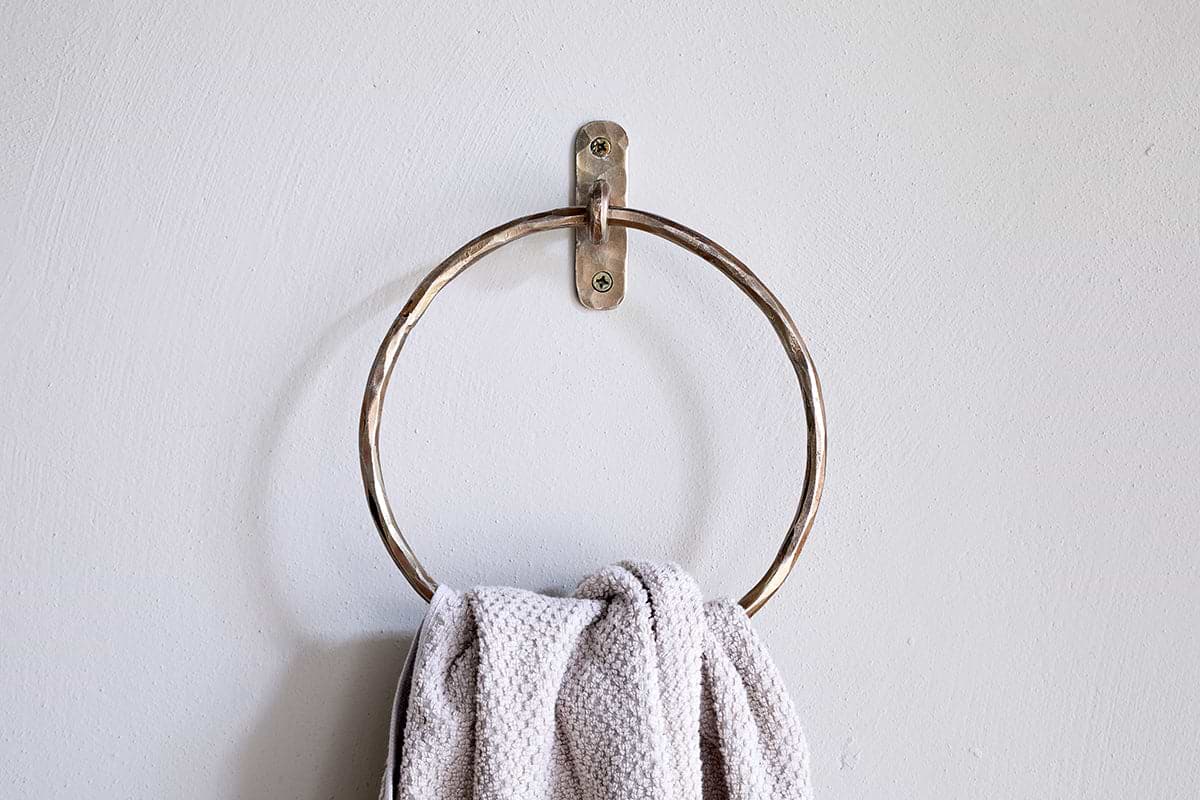 An Olani Towel Ring - Antique Brass by nkuku is mounted on a plain white wall, with a textured light gray hand towel draped over it, adding an elegant and minimal touch to the bathroom decor.