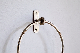 A close-up of the Olani Towel Ring - Antique Brass by nkuku, showing its textured, hammered surface and oval backplate with two visible screws, mounted on a pale grey wall as an elegant bathroom accessory.