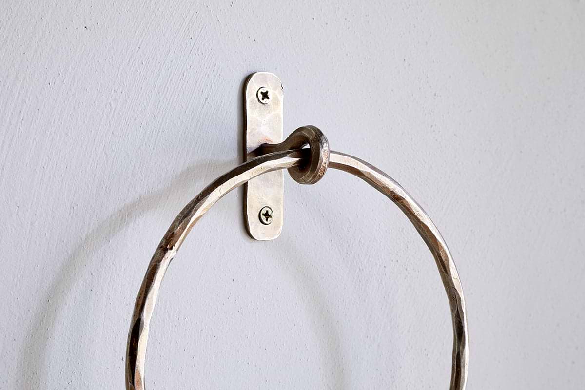 A close-up of the Olani Towel Ring - Antique Brass by nkuku, showing its textured, hammered surface and oval backplate with two visible screws, mounted on a pale grey wall as an elegant bathroom accessory.