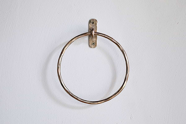 The Olani Towel Ring - Antique Brass by nkuku, a round bathroom accessory with a slightly twisted texture, is mounted on a plain white wall using a small metal bracket and two visible screws, with no towels hanging from it.