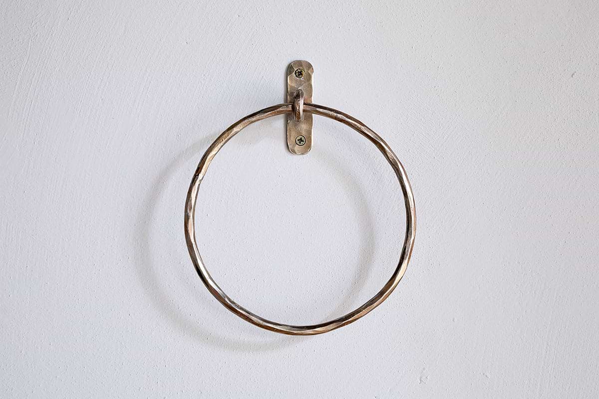 The Olani Towel Ring - Antique Brass by nkuku, a round bathroom accessory with a slightly twisted texture, is mounted on a plain white wall using a small metal bracket and two visible screws, with no towels hanging from it.