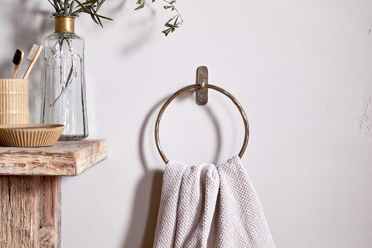 An Olani Towel Ring - Antique Brass by nkuku, holding a textured white towel, is mounted on a white wall. Nearby rustic shelves display olive branches, toothbrushes, and a dish for a minimalist bathroom look highlighted by this sleek accessory.