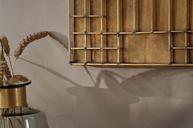 A decorative glass vase with dried grasses sits by a beige wall. Above, the Oni Printers Drawer - Brass by nkuku casts a shadow, its geometric design blending seamlessly with the neutral, warm color palette.