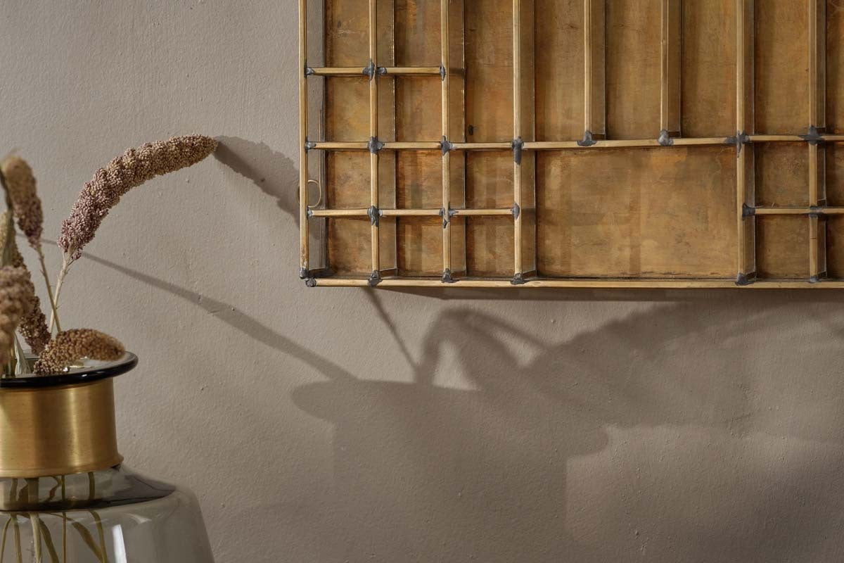 A decorative glass vase with dried grasses sits by a beige wall. Above, the Oni Printers Drawer - Brass by nkuku casts a shadow, its geometric design blending seamlessly with the neutral, warm color palette.