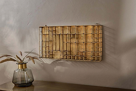 The Oni Printers Drawer - Brass by nkuku, a geometric gold wall sculpture inspired by traditional printing drawers, hangs on a light gray wall above wood. To the left, a gold-rimmed glass vase with wheat stems complements this unique wall art.