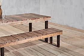 The nkuku Oso Wooden Bench, featuring rustic reclaimed wood and dark metal legs, sits next to a matching wooden table on a light floor against a plain gray wall. A glass bottle with dried plants decorates the table's corner.