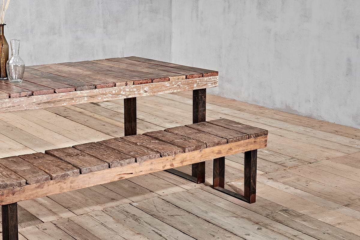 The nkuku Oso Wooden Bench, featuring rustic reclaimed wood and dark metal legs, sits next to a matching wooden table on a light floor against a plain gray wall. A glass bottle with dried plants decorates the table's corner.