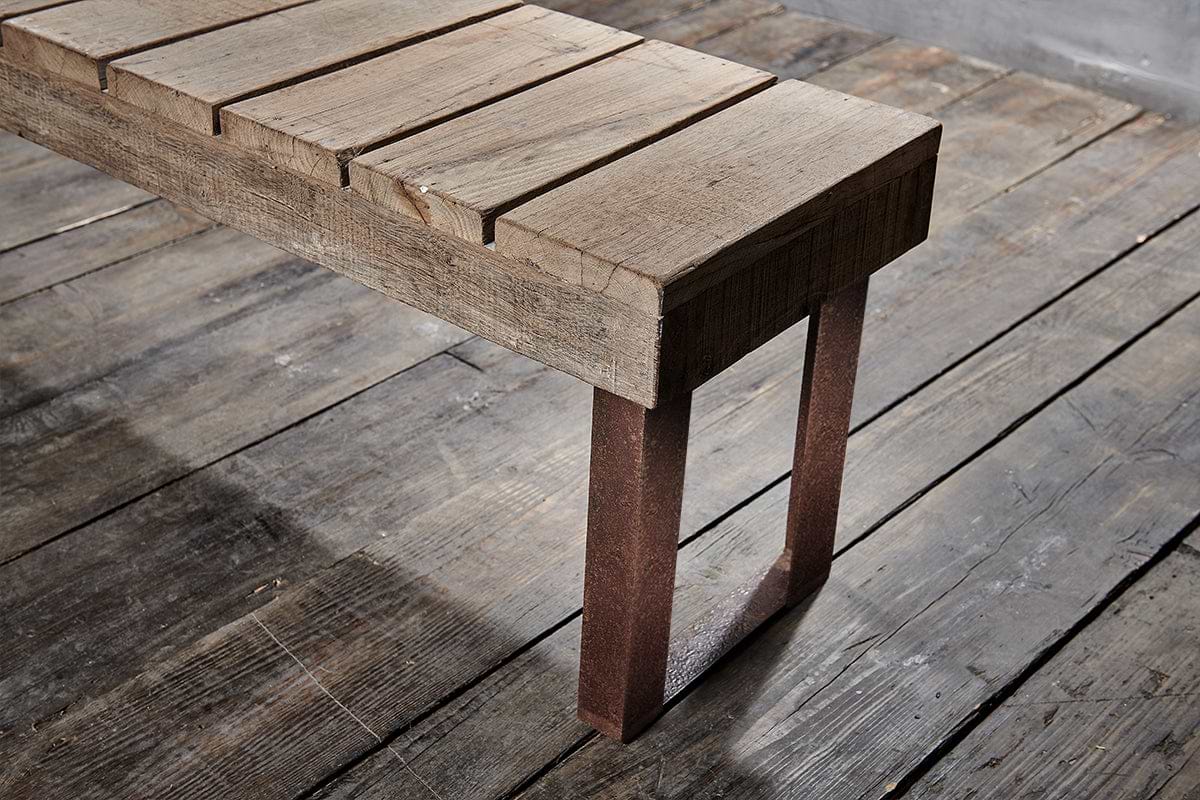 The Oso Wooden Bench by nkuku features a rustic slat design and sturdy metal legs, enhancing any gray-toned, industrial space with its aged charm and minimalist elegance.