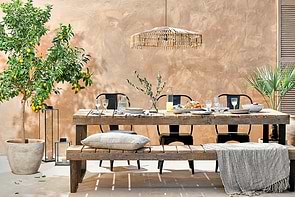 A rustic outdoor dining area showcases the nkuku Oso Wooden Dining Table set for a meal, paired with black chairs, a cushioned bench, potted lemon tree and palm, two lanterns, and a woven pendant light against a beige stucco wall.