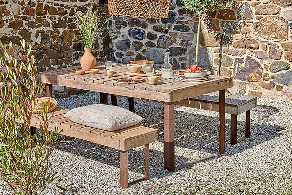 In a rustic outdoor setting, the nkuku Oso Wooden Dining Table with iron legs sits on gravel, topped with a flower vase, pottery, fruit plates, and cups. A pillow accents one bench amid a stone wall and potted plants for a cozy ambiance.