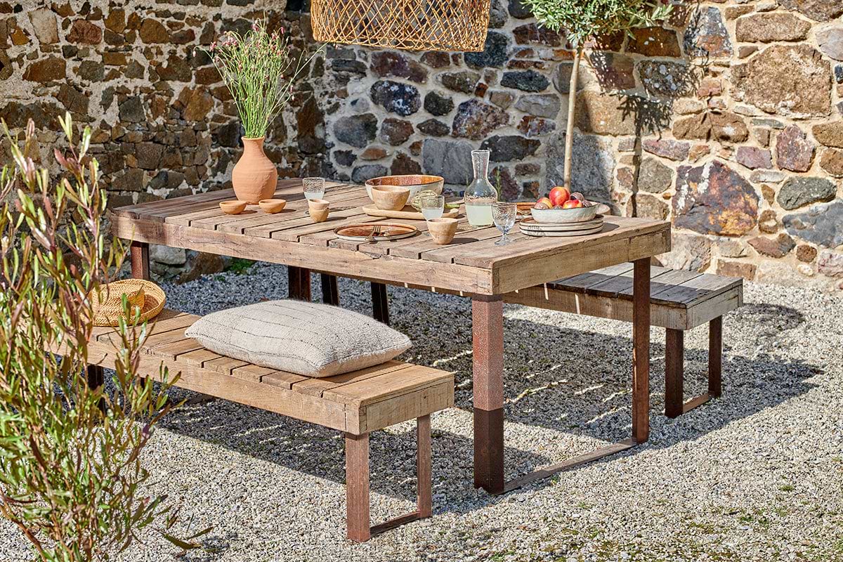In a rustic outdoor setting, the nkuku Oso Wooden Dining Table with iron legs sits on gravel, topped with a flower vase, pottery, fruit plates, and cups. A pillow accents one bench amid a stone wall and potted plants for a cozy ambiance.