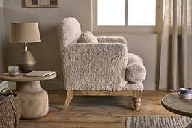The Otas Teddy Cotton Armchair by nkuku in off white sits on a wooden floor beside a rustic side table featuring a lamp and an open book.