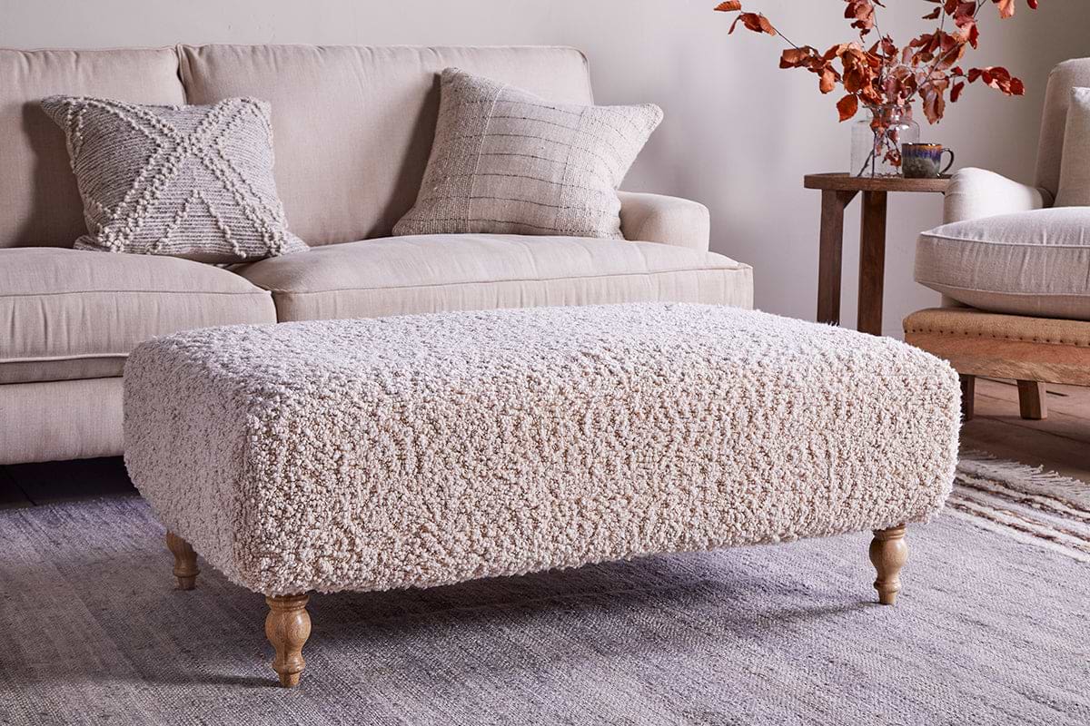The Otas Teddy Ottoman in off white by nkuku, featuring wooden legs, stands on a woven rug before a cream sofa with textured pillows; a side table holding a mug and autumn leaves completes the cozy, neutral decor.