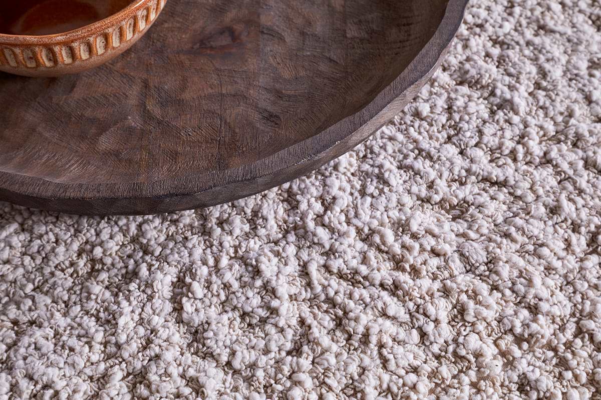 A close-up, angled shot shows a dark wooden tray and ceramic bowl resting on the nkuku Otas Teddy Ottoman - Off White, which features a mango wood base atop a plush, cream-colored shag rug.