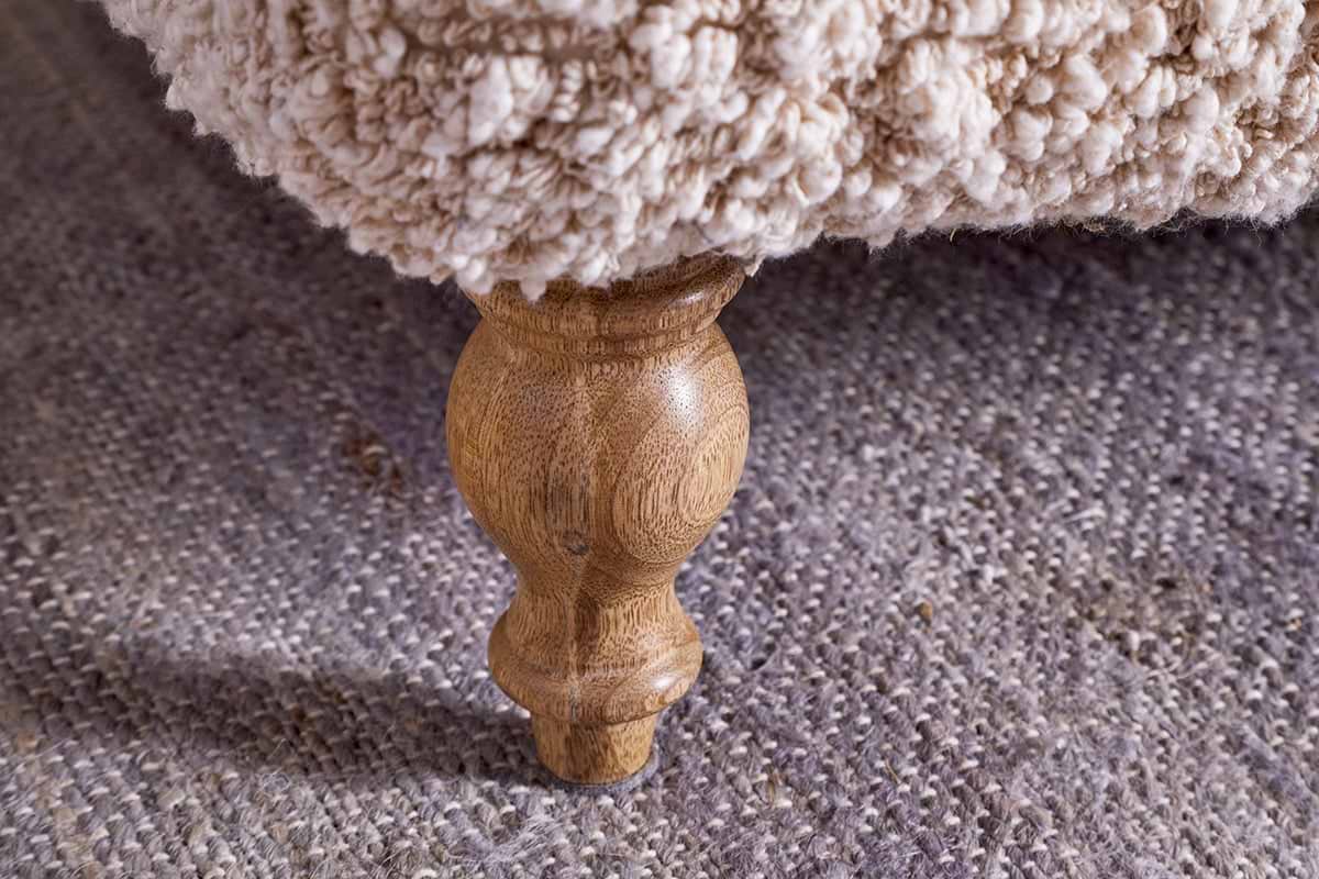 Close-up of a turned oak leg beneath nkuku’s Otas Teddy Ottoman - Off White. The cream boucle upholstery and textured grey rug accentuate the ottoman’s elegant, rounded form.