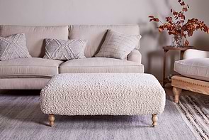 A cozy living room with a beige sofa, three textured pillows, a large nkuku Otas Teddy Ottoman in off white with wooden legs, a neutral rug, and a side table holding a glass vase of autumnal branches and books.