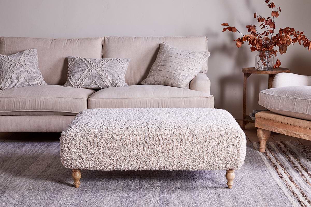 A cozy living room with a beige sofa, three textured pillows, a large nkuku Otas Teddy Ottoman in off white with wooden legs, a neutral rug, and a side table holding a glass vase of autumnal branches and books.