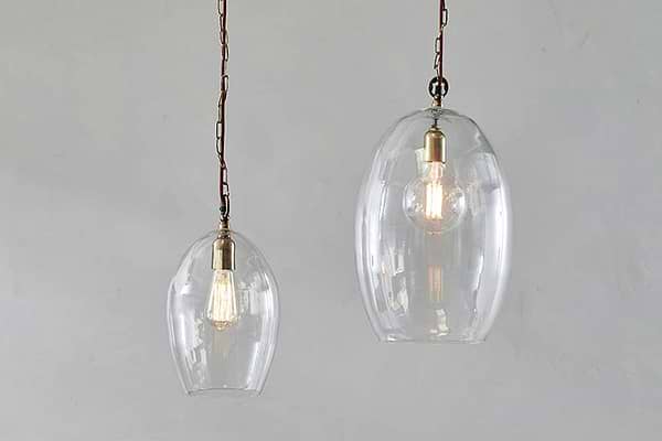 Two Otoro Recycled Glass Pendant - Clear - Small Oval lights by nkuku, with brass fittings and brown chains, hang against a light grey wall. The smaller pendant emits a warm glow, highlighting the bulbs and elegant recycled glass domes.