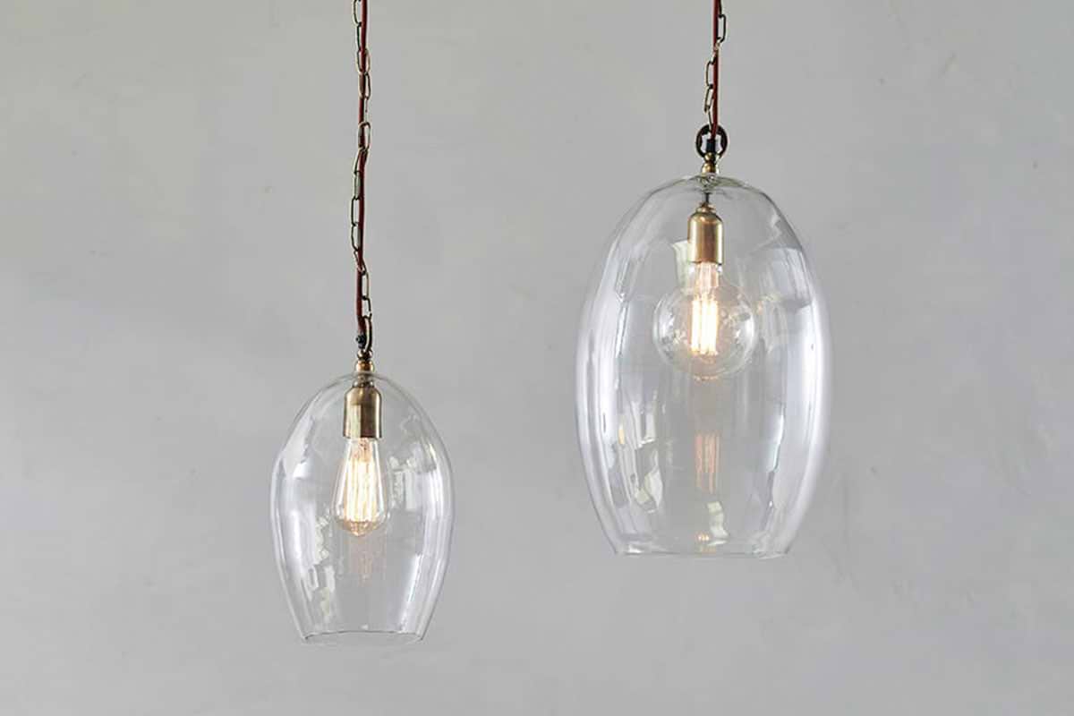 Two Otoro Recycled Glass Pendant - Clear - Small Oval lights by nkuku, with brass fittings and brown chains, hang against a light grey wall. The smaller pendant emits a warm glow, highlighting the bulbs and elegant recycled glass domes.